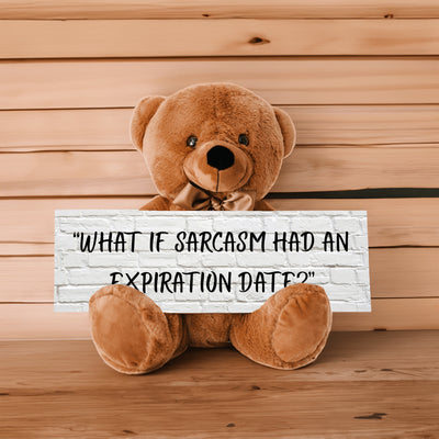 A teddy bear sitting on a wooden surface holding a sign with the phrase 'WHAT IF SARCASM HAD AN EXPIRATION DATE?'