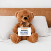 Brown teddy bear holding a sign on a bed
