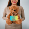 "Plush teddy bear holding a card with the message 'Mom, We Love You – Forever Our Guide' on a pink floral background."