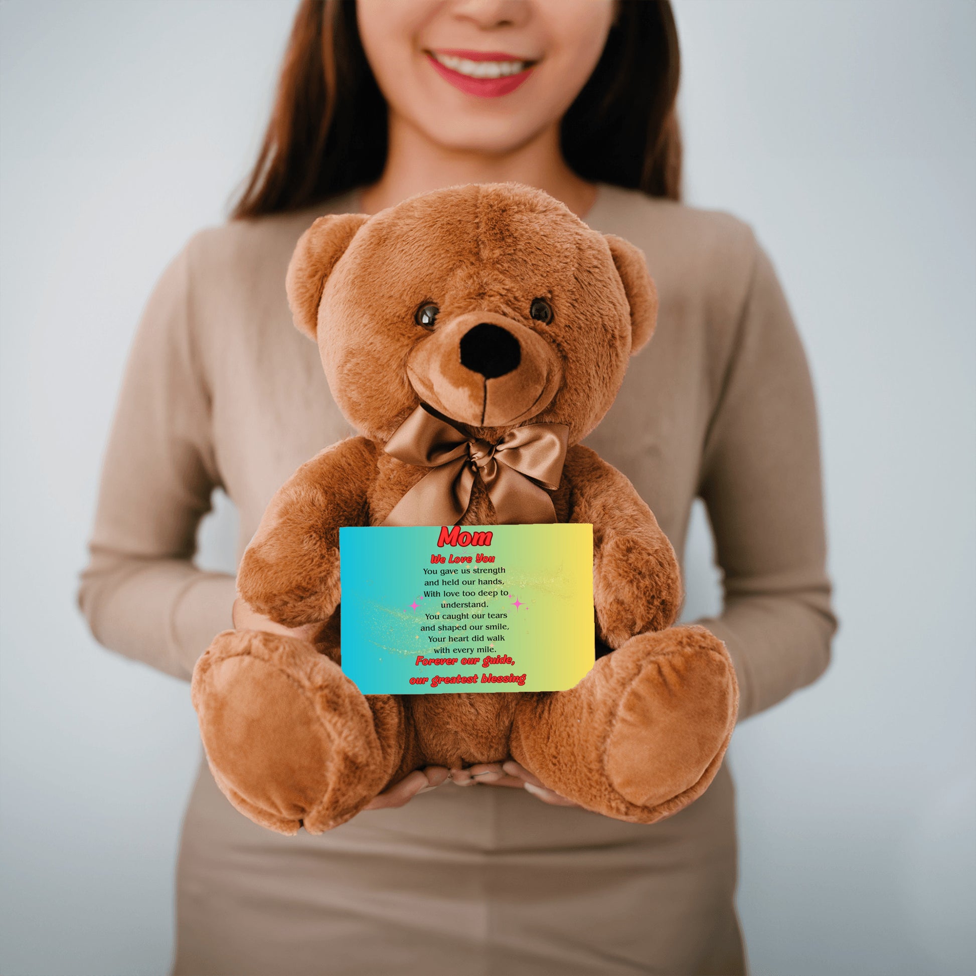 "Plush teddy bear holding a card with the message 'Mom, We Love You – Forever Our Guide' on a pink floral background."