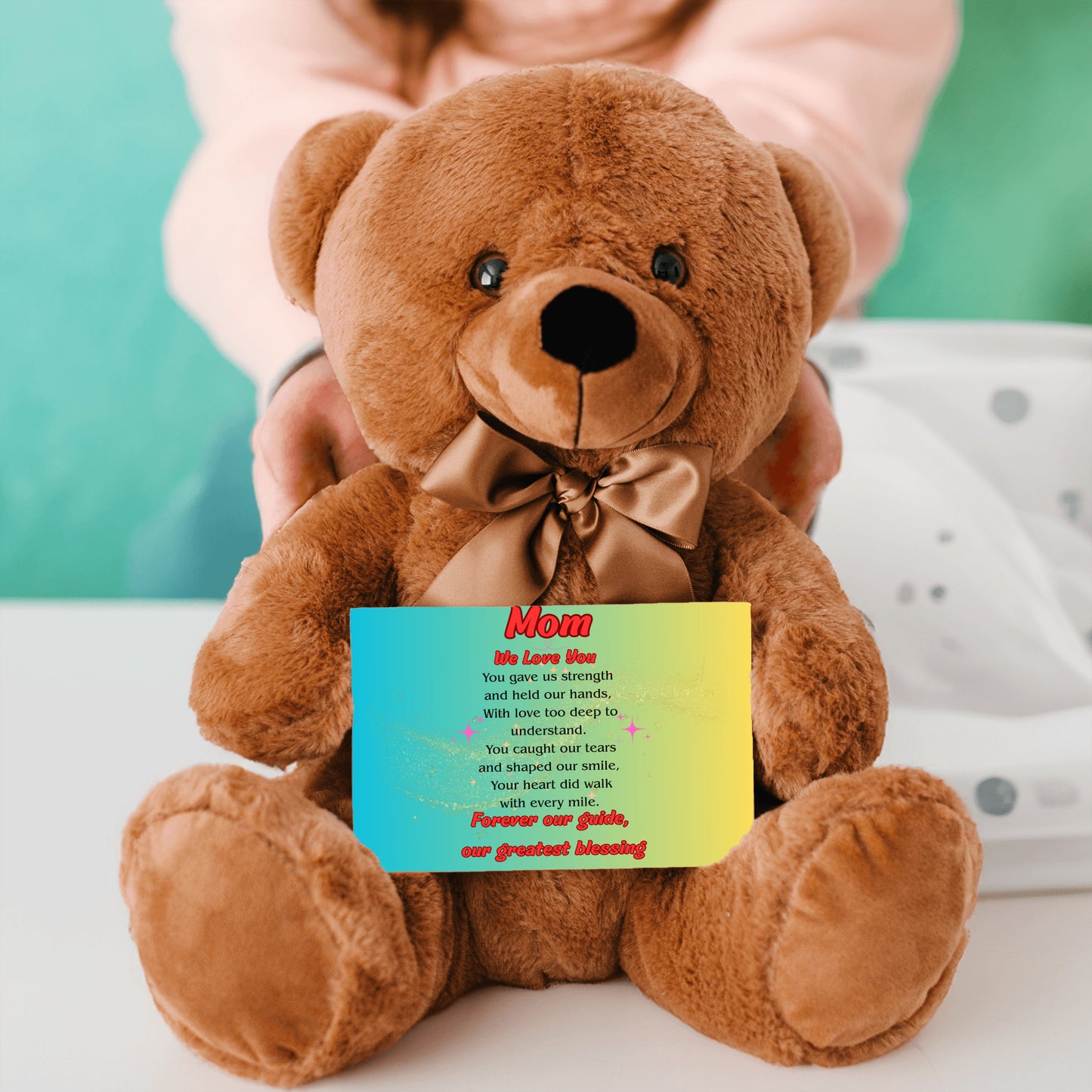 "Plush teddy bear holding a card with the message 'Mom, We Love You – Forever Our Guide' on a pink floral background."
