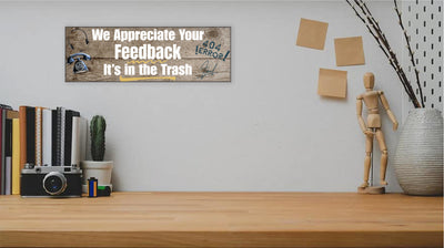 A rectangular wooden plaque featuring the sarcastic phrase "We Appreciate Your Feedback – It’s in the Trash" in bold white and red text on a dark vintage background with ornamental border details.