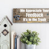 A rectangular wooden plaque featuring the sarcastic phrase "We Appreciate Your Feedback – It’s in the Trash" in bold white and red text on a dark vintage background with ornamental border details.