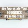 A rectangular wooden plaque featuring the sarcastic phrase "We Appreciate Your Feedback – It’s in the Trash" in bold white and red text on a dark vintage background with ornamental border details.