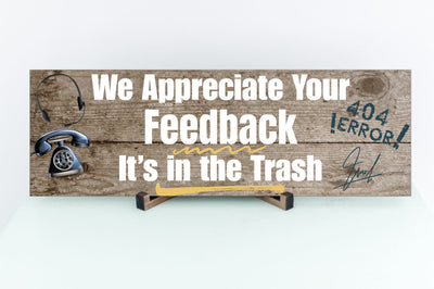 A rectangular wooden plaque featuring the sarcastic phrase "We Appreciate Your Feedback – It’s in the Trash" in bold white and red text on a dark vintage background with ornamental border details.