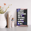 A decorative wood plaque with bold, distressed text reading “It Won’t Happen Overnight,” featuring a minimalist style and comic flair.