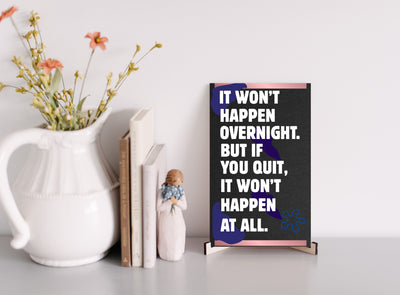 A decorative wood plaque with bold, distressed text reading “It Won’t Happen Overnight,” featuring a minimalist style and comic flair.