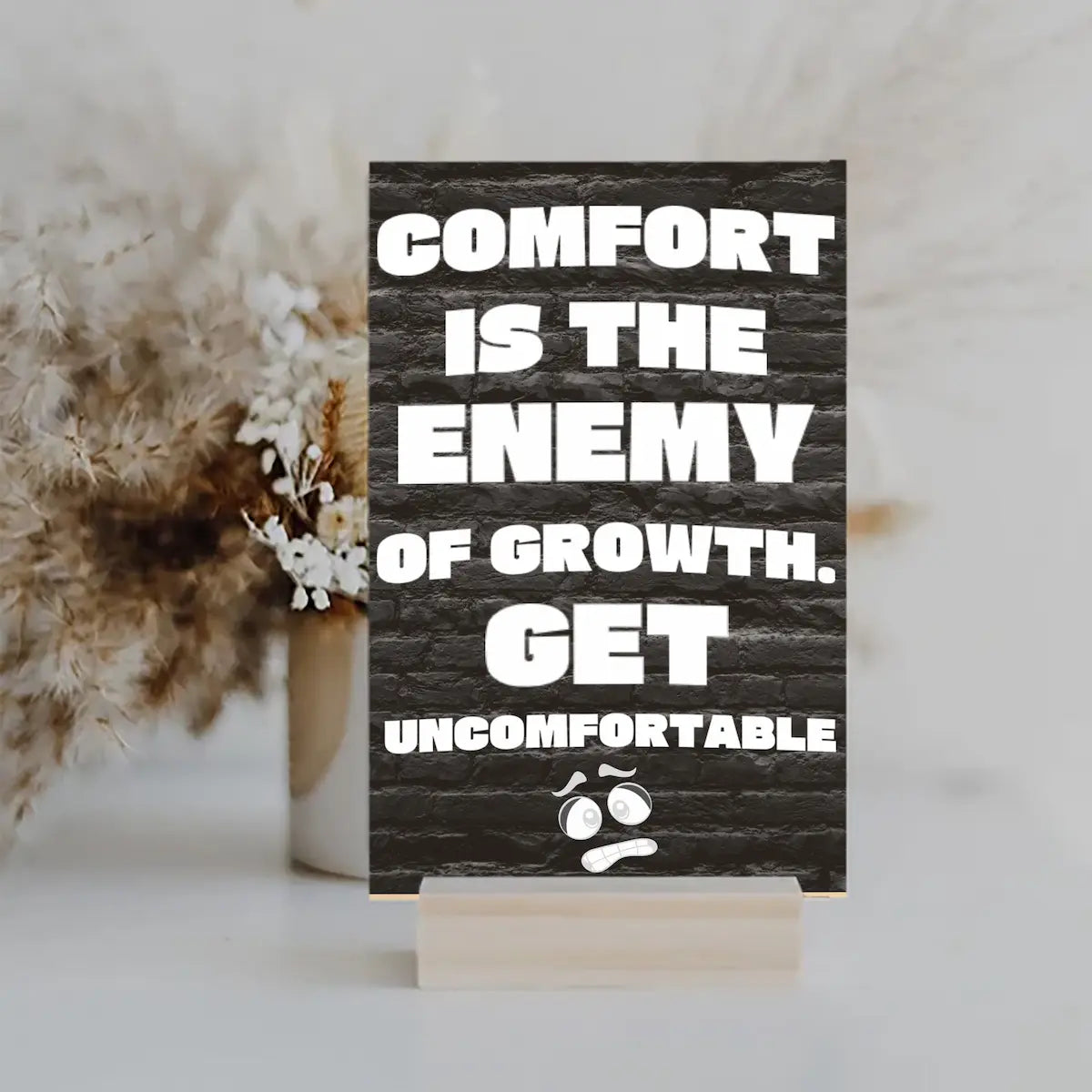 A modern wall plaque that reads “Comfort Is the Enemy of Growth” in bold, minimalist black letters on a clean woodgrain background.