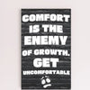 A modern wall plaque that reads “Comfort Is the Enemy of Growth” in bold, minimalist black letters on a clean woodgrain background.