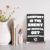 A modern wall plaque that reads “Comfort Is the Enemy of Growth” in bold, minimalist black letters on a clean woodgrain background.