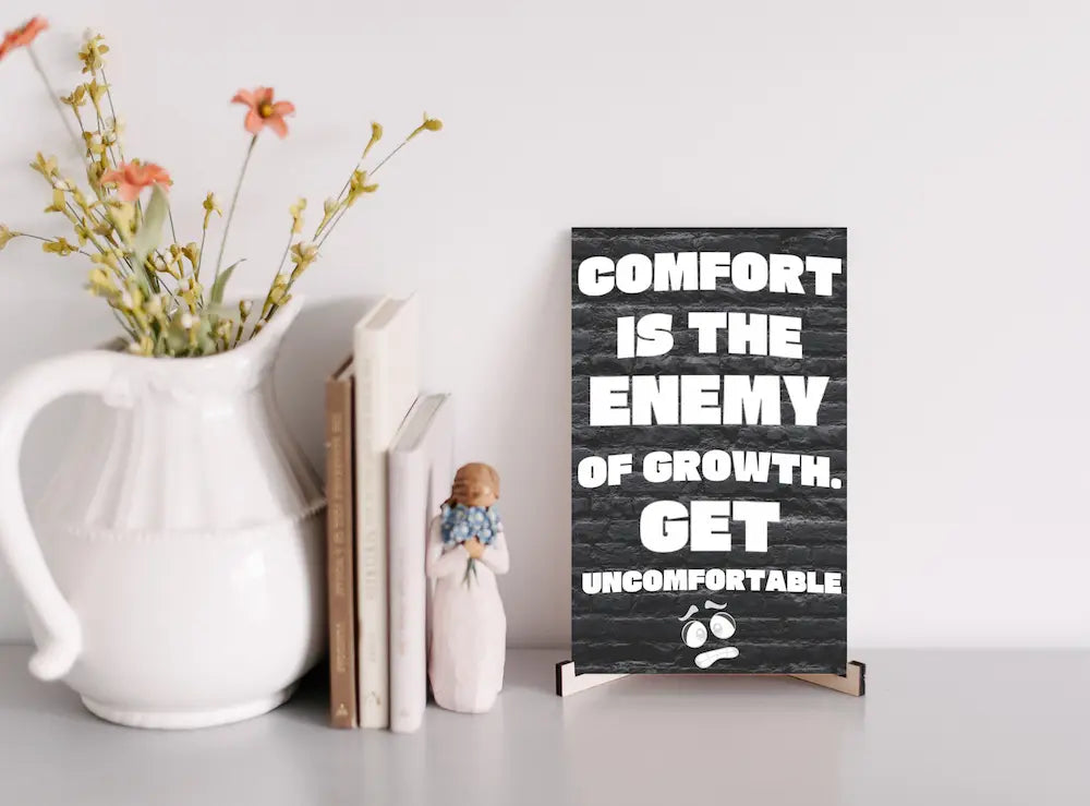 A modern wall plaque that reads “Comfort Is the Enemy of Growth” in bold, minimalist black letters on a clean woodgrain background.