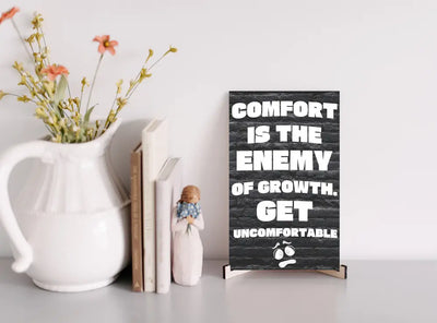 A modern wall plaque that reads “Comfort Is the Enemy of Growth” in bold, minimalist black letters on a clean woodgrain background.