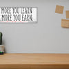 A modern motivational plaque with bold block text that reads “The More You Learn – The More You Earn” on a clean woodgrain background.