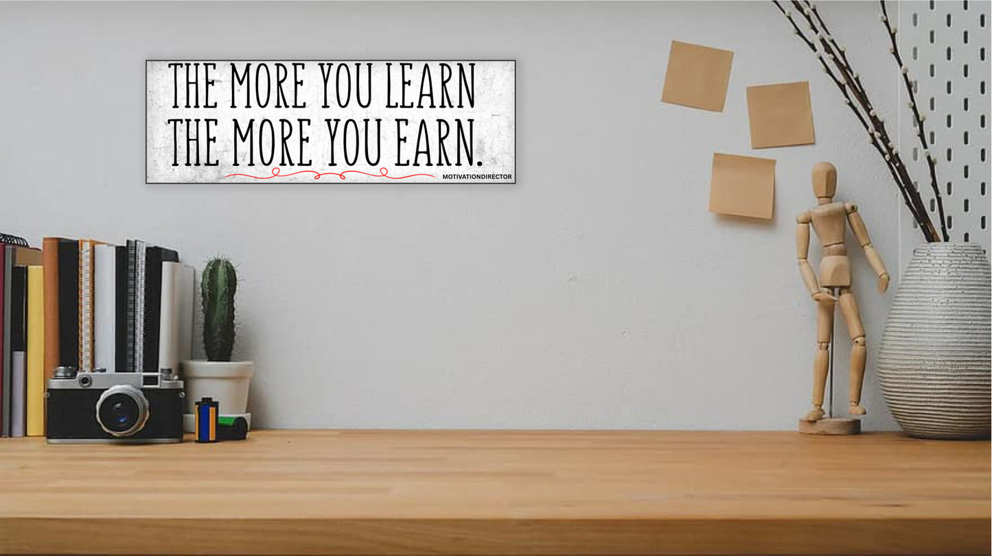 A modern motivational plaque with bold block text that reads “The More You Learn – The More You Earn” on a clean woodgrain background.