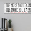 A modern motivational plaque with bold block text that reads “The More You Learn – The More You Earn” on a clean woodgrain background.
