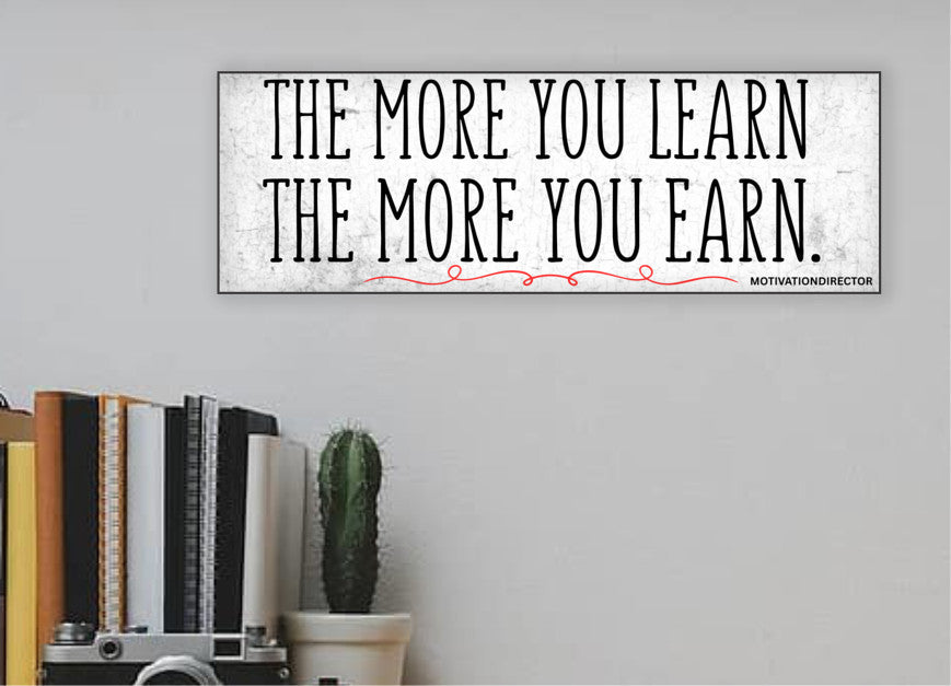 A modern motivational plaque with bold block text that reads “The More You Learn – The More You Earn” on a clean woodgrain background.