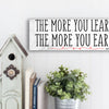A modern motivational plaque with bold block text that reads “The More You Learn – The More You Earn” on a clean woodgrain background.