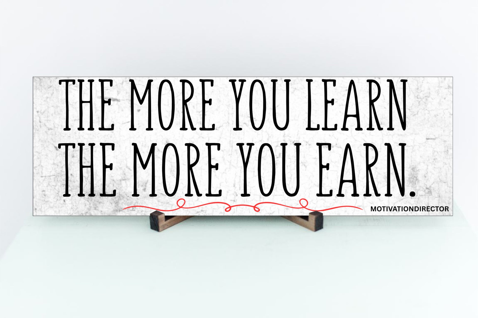 A modern motivational plaque with bold block text that reads “The More You Learn – The More You Earn” on a clean woodgrain background.