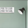 A modern motivational plaque with bold block text that reads “The More You Learn – The More You Earn” on a clean woodgrain background.