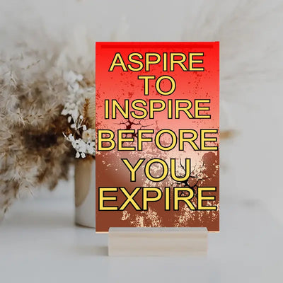 Modern inspirational plaque with the quote “Aspire to Inspire Before You Expire” in bold, artistic typography on wood, styled on a workspace wall.