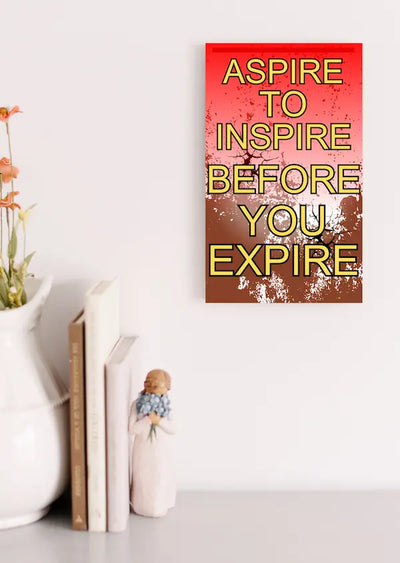 Modern inspirational plaque with the quote “Aspire to Inspire Before You Expire” in bold, artistic typography on wood, styled on a workspace wall.