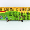 Wooden plaque with bold pink lettering reading “Cart Girl Magic In Progress” featuring cartoon-style golf cart, drink tray, and dollar signs—displayed outdoors.
