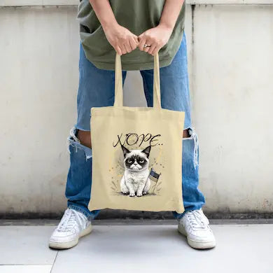 A beige canvas tote bag featuring a grumpy cartoon cat with the word “NOPE” in bold block letters.