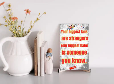 Wooden plaque with bold text: “Your biggest fans are strangers. Your biggest hater is someone you know,” styled with comic-style humor.