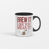 White ceramic coffee mug with bold black text reading "Brew It Like You Mean It" sitting on a wooden desk with steam rising from the cup.