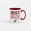 White ceramic coffee mug with bold black text reading "Brew It Like You Mean It" sitting on a wooden desk with steam rising from the cup.