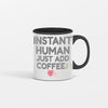 White ceramic coffee mug with bold text reading "INSTANT HUMAN, JUST ADD COFFEE" in black lettering, placed on a rustic wooden table with spilled coffee beans and steam rising.