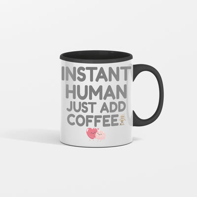 White ceramic coffee mug with bold text reading "INSTANT HUMAN, JUST ADD COFFEE" in black lettering, placed on a rustic wooden table with spilled coffee beans and steam rising.