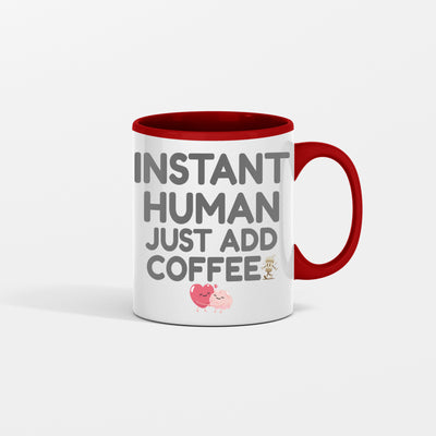 White ceramic coffee mug with bold text reading "INSTANT HUMAN, JUST ADD COFFEE" in black lettering, placed on a rustic wooden table with spilled coffee beans and steam rising.