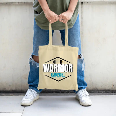 Cinematic shot of the Warrior Blueprint Tote Bag resting beside gym gear and a water bottle, morning light hitting the bold logo, symbolizing strength, motion, and warrior focus.