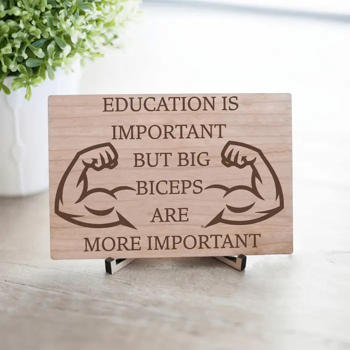 Wooden plaque reading ‘Education is Important but Bigger Biceps Are More Important,’ displayed on gym wall next to dumbbells, sarcastic motivational décor.
