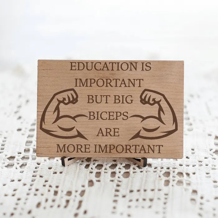 Wooden plaque reading ‘Education is Important but Bigger Biceps Are More Important,’ displayed on gym wall next to dumbbells, sarcastic motivational décor.