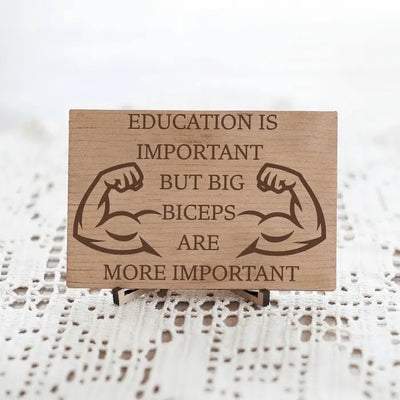 Wooden plaque reading ‘Education is Important but Bigger Biceps Are More Important,’ displayed on gym wall next to dumbbells, sarcastic motivational décor.