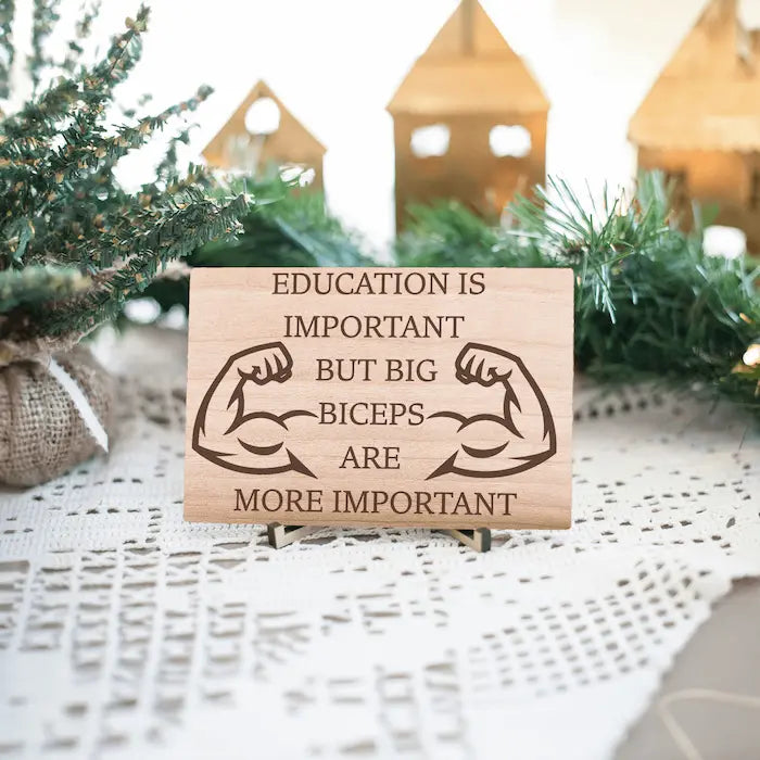 Wooden plaque reading ‘Education is Important but Bigger Biceps Are More Important,’ displayed on gym wall next to dumbbells, sarcastic motivational décor.