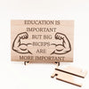Wooden plaque reading ‘Education is Important but Bigger Biceps Are More Important,’ displayed on gym wall next to dumbbells, sarcastic motivational décor.