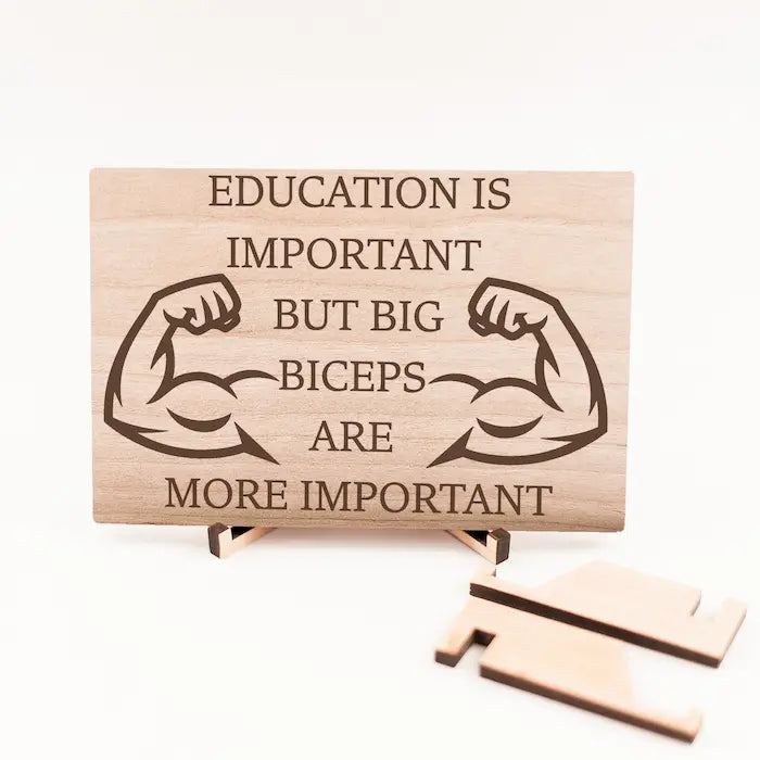 Wooden plaque reading ‘Education is Important but Bigger Biceps Are More Important,’ displayed on gym wall next to dumbbells, sarcastic motivational décor.