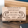 Wooden plaque reading ‘Education is Important but Bigger Biceps Are More Important,’ displayed on gym wall next to dumbbells, sarcastic motivational décor.
