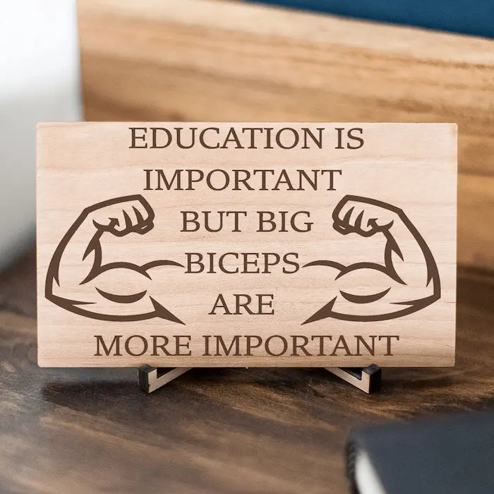 Wooden plaque reading ‘Education is Important but Bigger Biceps Are More Important,’ displayed on gym wall next to dumbbells, sarcastic motivational décor.