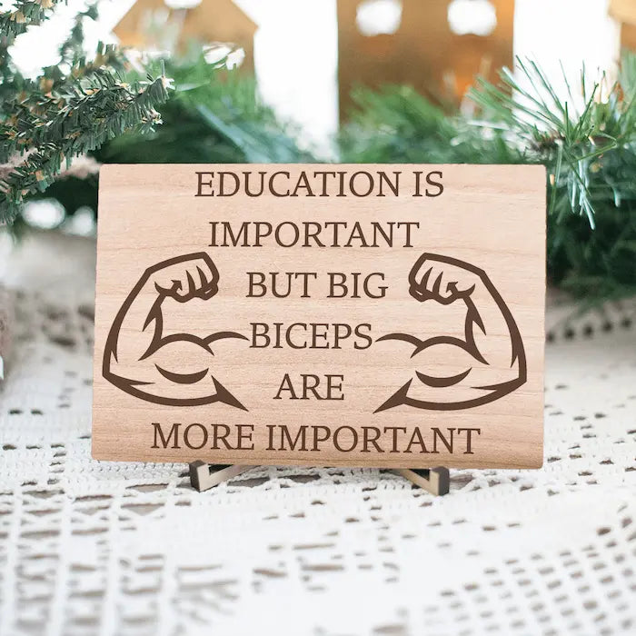 Wooden plaque reading ‘Education is Important but Bigger Biceps Are More Important,’ displayed on gym wall next to dumbbells, sarcastic motivational décor.