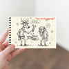 Hand holding a small notebook with a Christmas-themed illustration of a reindeer and person, on a neutral background.